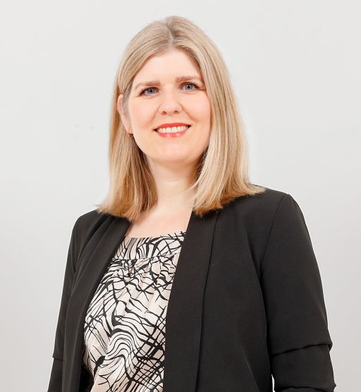 Frau mit blonden Haaren und rotem Lippenstift, schwarzem Blazer über gemusterter Bluse, lächelt vor hellgrauem Hintergrund.