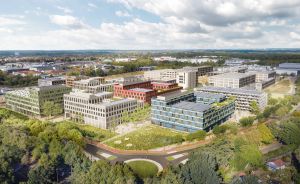 Luftaufnahme eines modernen Gebäudekomplexes mit farbigen Fassaden, umgeben von Bäumen und Grünflächen in einer Stadt.
