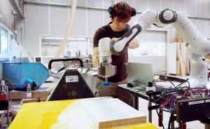 Mann mit Gehörschutz arbeitet in Werkstatt mit weißem Roboterarm an einem Werkstück auf gelbem Tisch.
