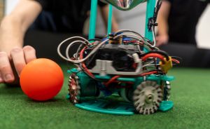 Türkiser Roboter mit Rädern und Elektronik auf grünem Untergrund, daneben orangefarbener Ball und menschliche Hand.