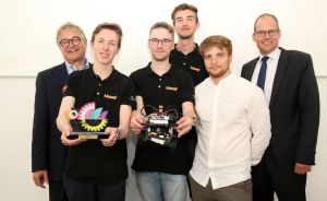 Gruppe von 6 Männern, zwei halten Roboter und Trophäe, vier in schwarzen Polo-Shirts, zwei in Businesskleidung.