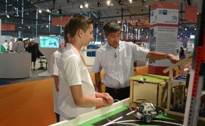 Ein Mann und ein Jugendlicher stehen an einem Tisch mit einem Roboter auf einer Spielfläche in einer Messehalle.