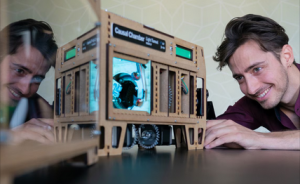 Mann schaut lächelnd auf ein beleuchtetes Gerät mit Aufschrift „Cloud Chamber" aus Holz; Spiegelbild links sichtbar.