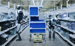 Zwei humanoide Roboter stehen in einem Lagergang zwischen Regalen, dahinter ein fahrerloses Transportfahrzeug mit blauem Regal.
