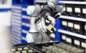 Roboterarm mit mechanischer Greifhand hält ein kleines Bauteil über einer Ablage mit sortierten Komponenten.