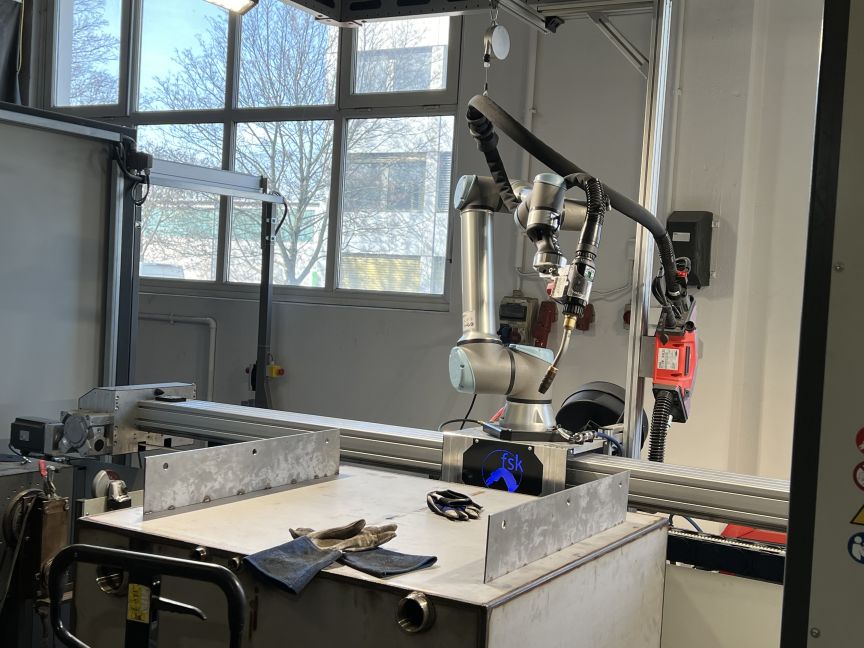 Industrieroboterarm auf einem Arbeitstisch in einer Werkstatt, daneben Werkzeuge und Schutzhandschuhe.
