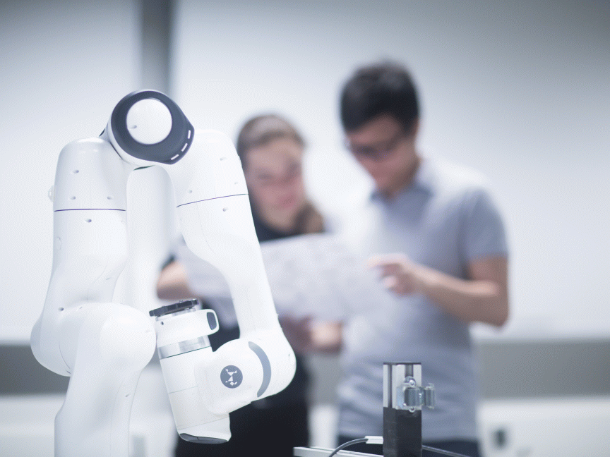 „Weißer Roboterarm im Vordergrund, im unscharfen Hintergrund zwei Personen, die ein Dokument betrachten."