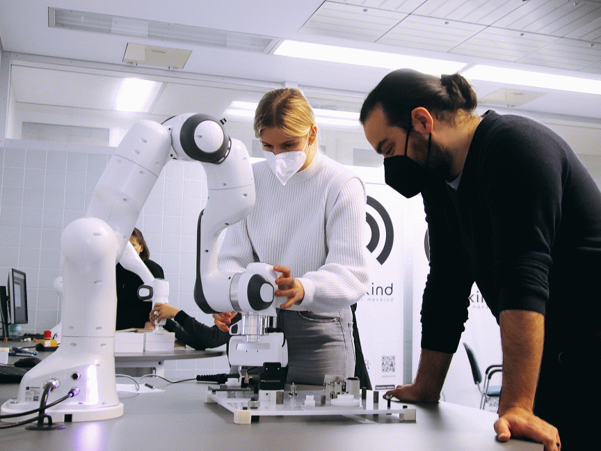 Zwei Personen mit Masken arbeiten an einem weißen Roboterarm auf einem Labortisch in einem hellen Raum.