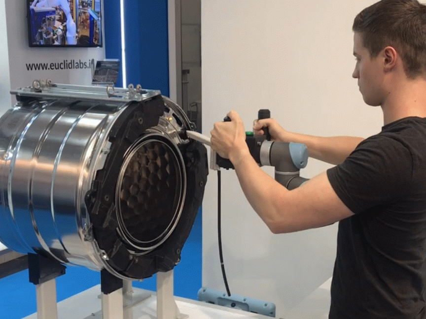 Mann in schwarzem T-Shirt bedient Roboterarm an zylindrischem Metallobjekt; Website „euclidlabs" im Hintergrund sichtbar.