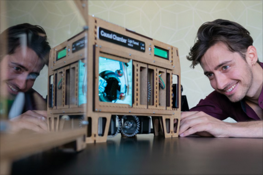 Mann schaut lächelnd auf ein beleuchtetes Gerät mit Aufschrift „Cloud Chamber" aus Holz; Spiegelbild links sichtbar.
