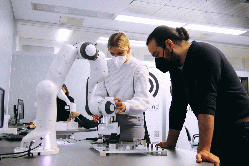 Zwei Personen mit Masken arbeiten an einem weißen Roboterarm auf einem Labortisch in einem hellen Raum.